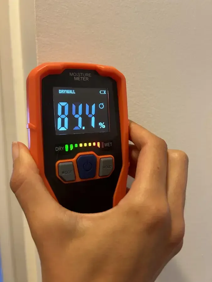 A handheld moisture meter displays a reading against a painted interior wall