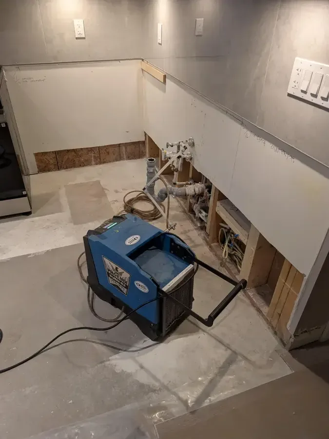 A blue air mover dries an opened wall base beside a tiled floor