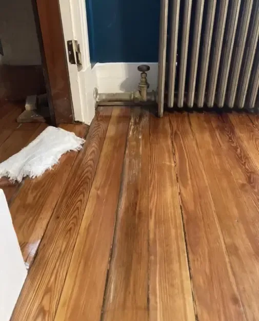 Wood floorboards meet a radiator and a wall with damaged base trim