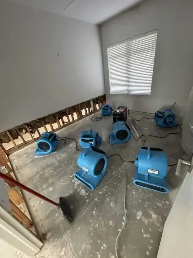 Blue air movers dry a bare floor beside opened wall bases