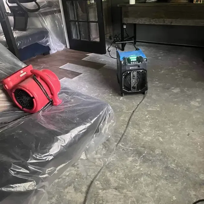 An air mover and dehumidifier stand beside furniture covered with protective plastic