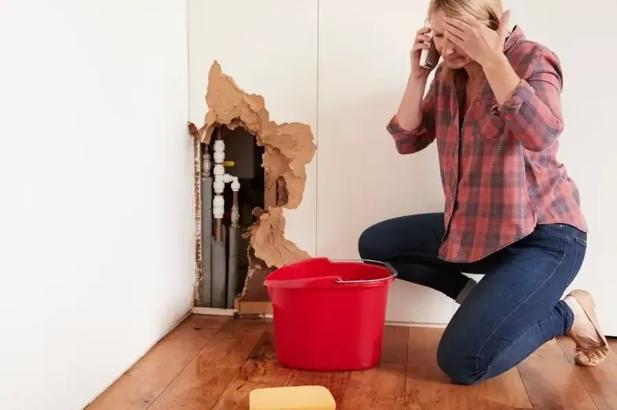 An open wall exposes pipes beside a red bucket on a wet floor