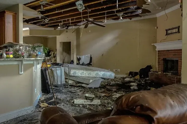 A collapsed ceiling exposes framing above debris and damaged furnishings in a living room