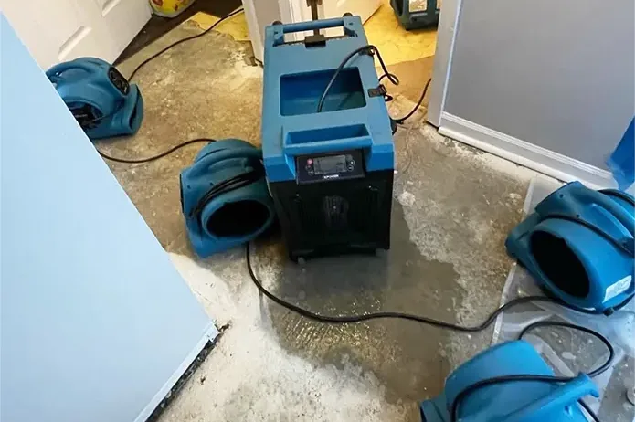 Blue air movers and a dehumidifier dry an exposed concrete floor beside opened walls